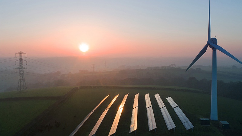 Landscape image featuring a wind turbine, multiple rows of solar panels, and a power pole.