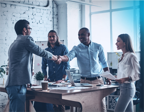 Two men and a woman are standing opposite another man in an office. Between them is a table over which two of the men are shaking hands.