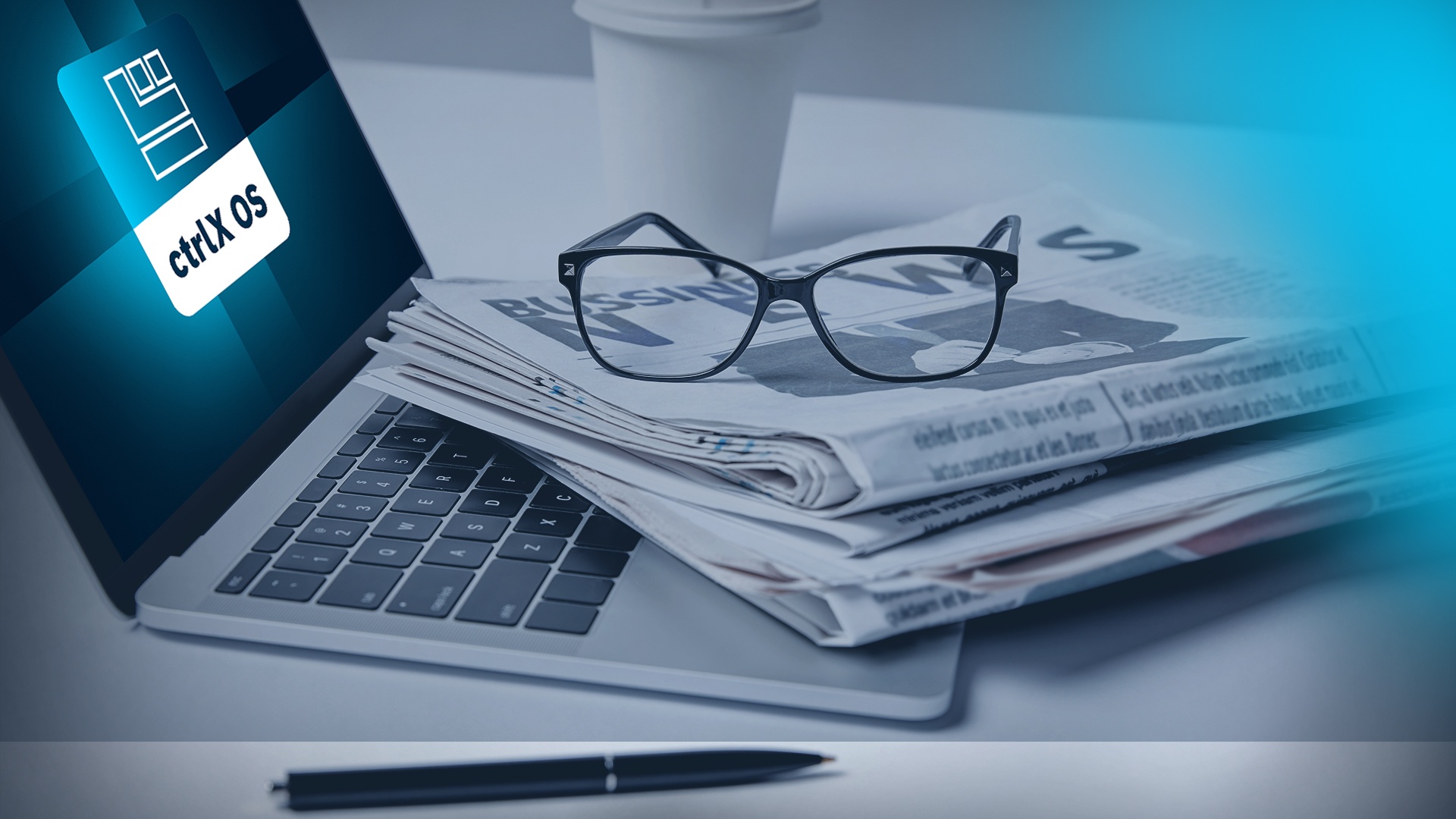 Table with a laptop viewed at an angle from the side, displaying the ctrlX OS app icon. A newspaper and a pair of black glasses are placed on the table.