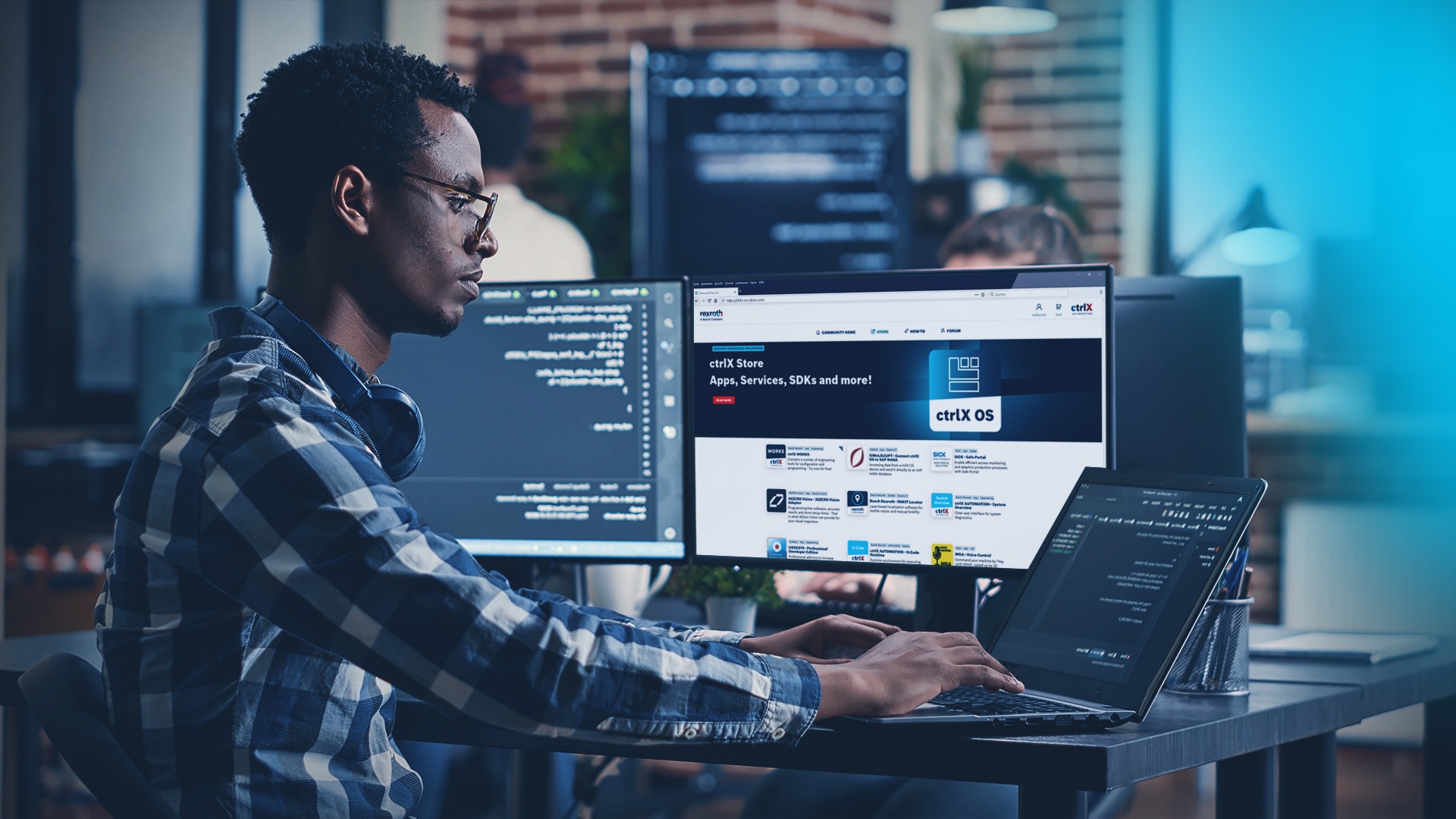 A man sits at a desk with two monitors and a laptop. One screen displays code, while the other shows the ctrlX OS Store website